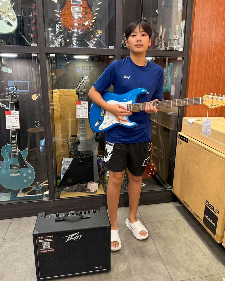 Teen in a guitar shop posing with a blue Stratocaster-style electric guitar; a Peavey amplifier sits on the floor beside them.