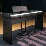 Black digital piano on a stand with three pedals, on a stage rug in a room with a plant in the background. ขายราคาพิเศษ