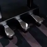 Close-up of three shiny metal piano pedals under a black piano on a dark striped carpet. ขายราคาพิเศษ