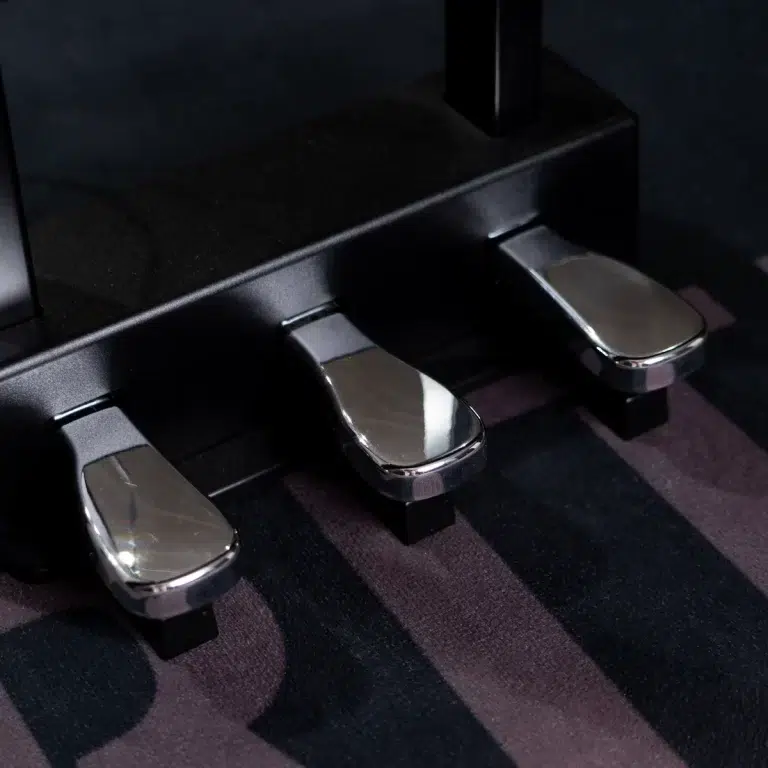 Close-up of three shiny metal piano pedals under a black piano on a dark striped carpet. ขายราคาพิเศษ