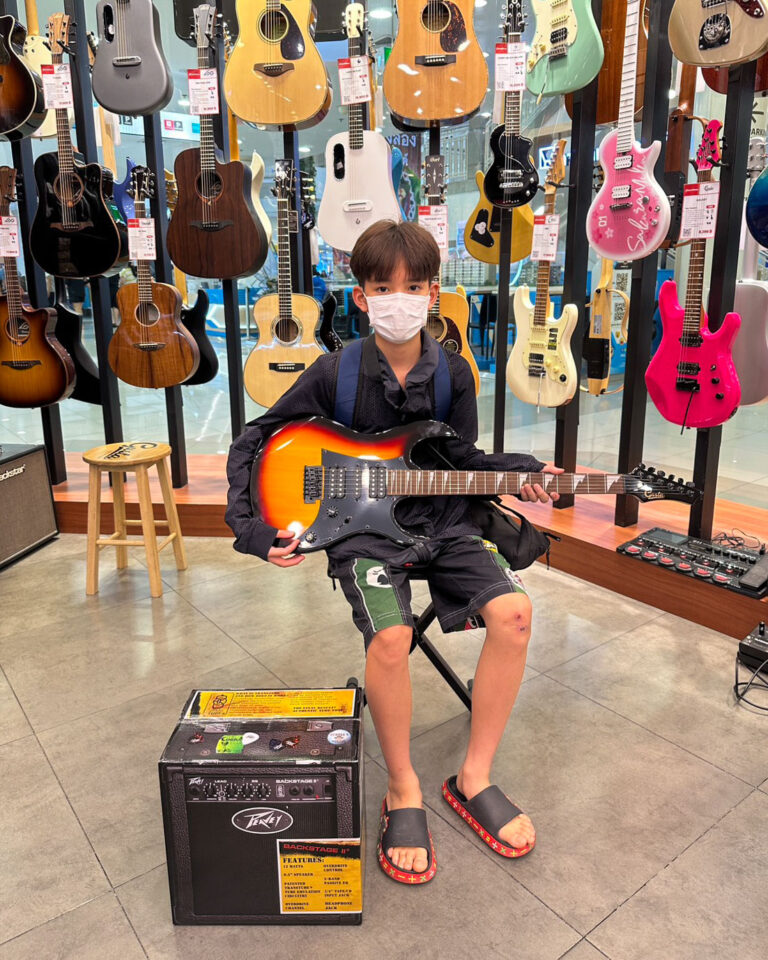 Child wearing a mask sits in a guitar shop, holding a sunburst electric guitar in front of a wall of hanging guitars.