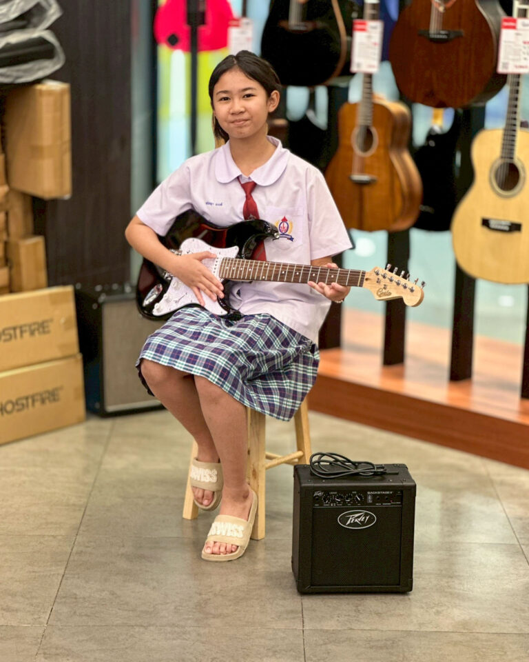 Young girl in a white school uniform and red tie sits on a stool, playing a black electric guitar in a music store.