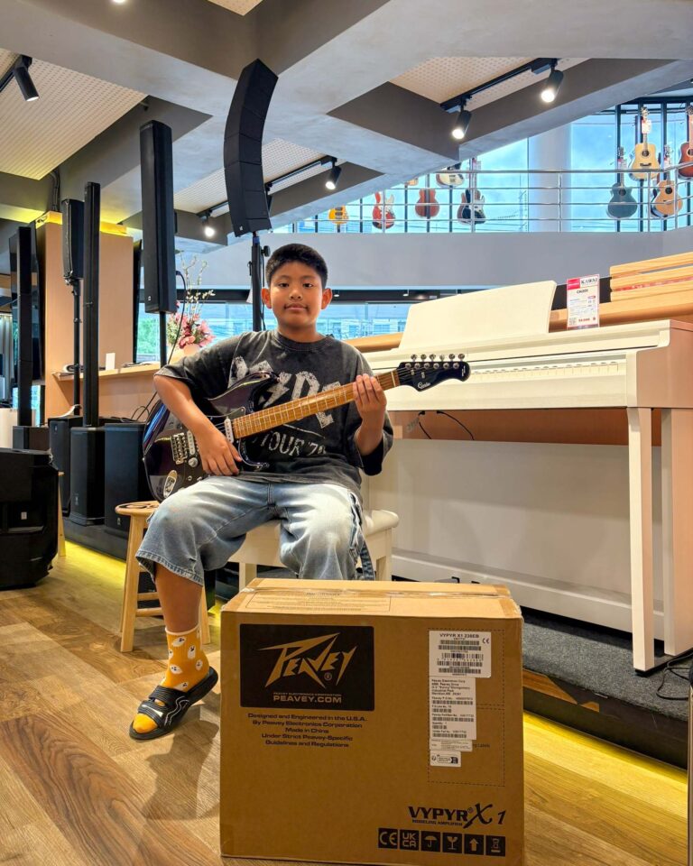 Young boy sitting on a stool, holding a black electric guitar in a music store, with guitars on the wall and a large Peavey box in front of him.