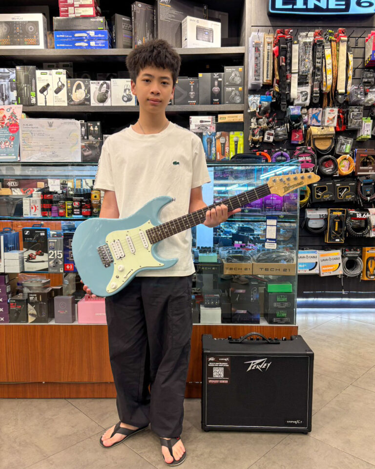 Young man in a store holding a light blue electric guitar in front of shelves of audio gear and accessories, with a Peavey amplifier nearby.