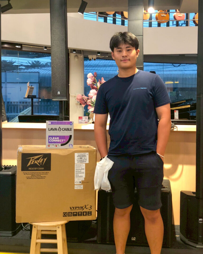 Young man in a navy shirt stands beside large cardboard boxes in a showroom with speakers and equipment around him.