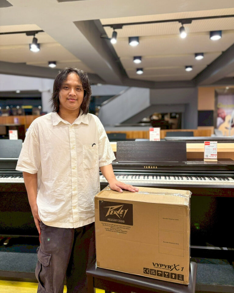 Man with shoulder-length hair and a light shirt stands beside a cardboard box in a music store, with a Yamaha keyboard behind him.