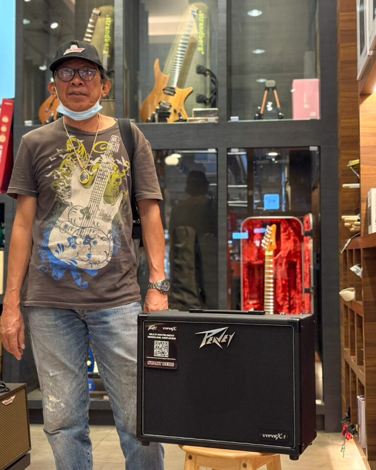 Man in a graphic tee and jeans stands in a guitar shop beside a Peavey amplifier on a wooden stool, with guitar displays behind him.
