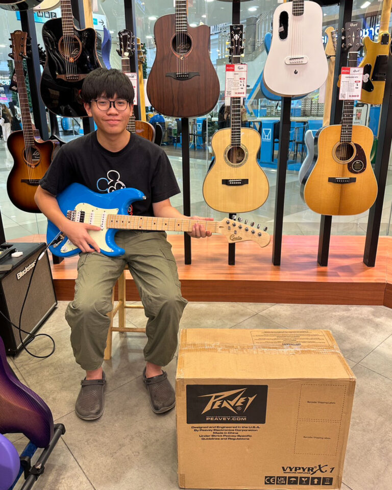 Young man sits on a stool in a guitar shop, playing a bright blue electric guitar with many acoustic guitars on racks behind him.