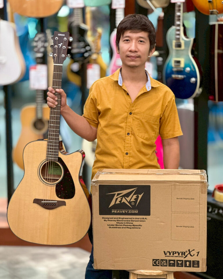 Man in a mustard shirt holding an acoustic guitar beside a large Peavey box in a guitar shop.