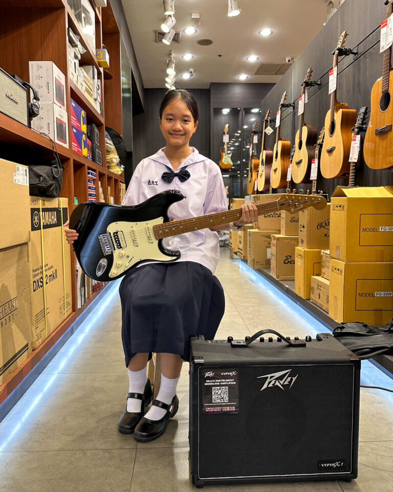 Girl in school uniform holding a blue electric guitar in a music store showroom with guitars on the wall and boxes on shelves behind her.