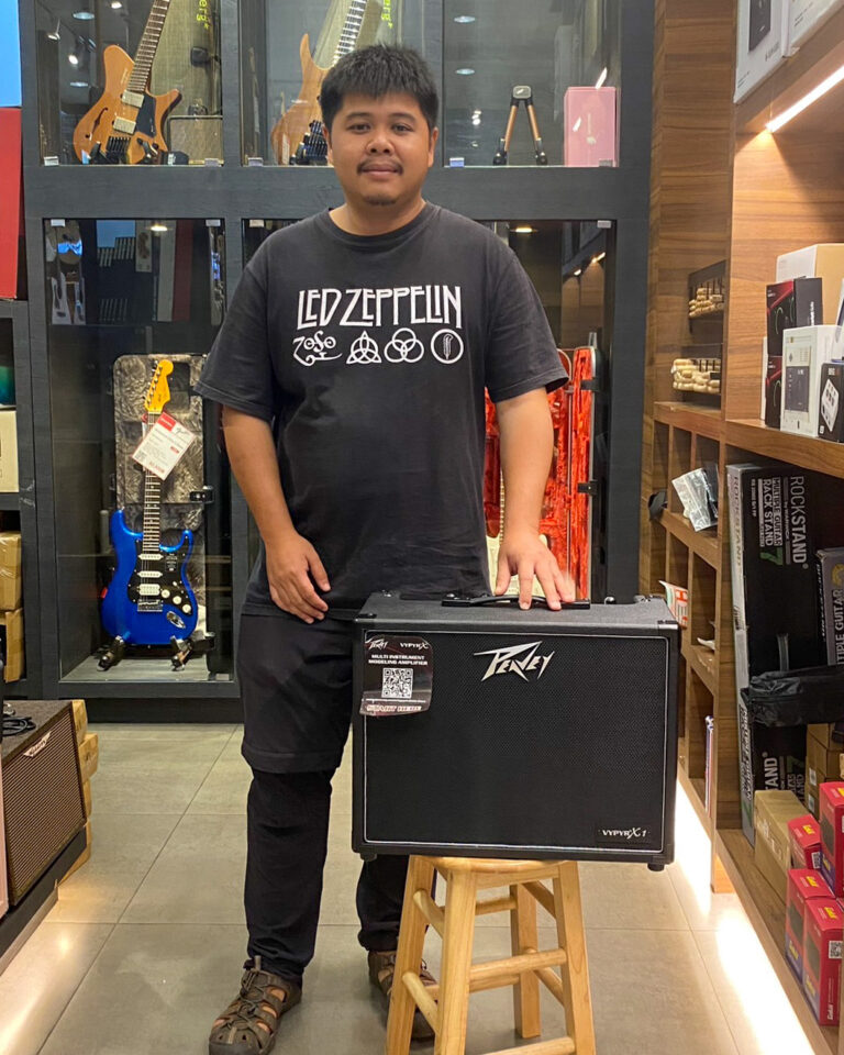 Man in a music store posing with a black Peavey guitar amplifier on a wooden stool in front of display cases with guitars behind him.