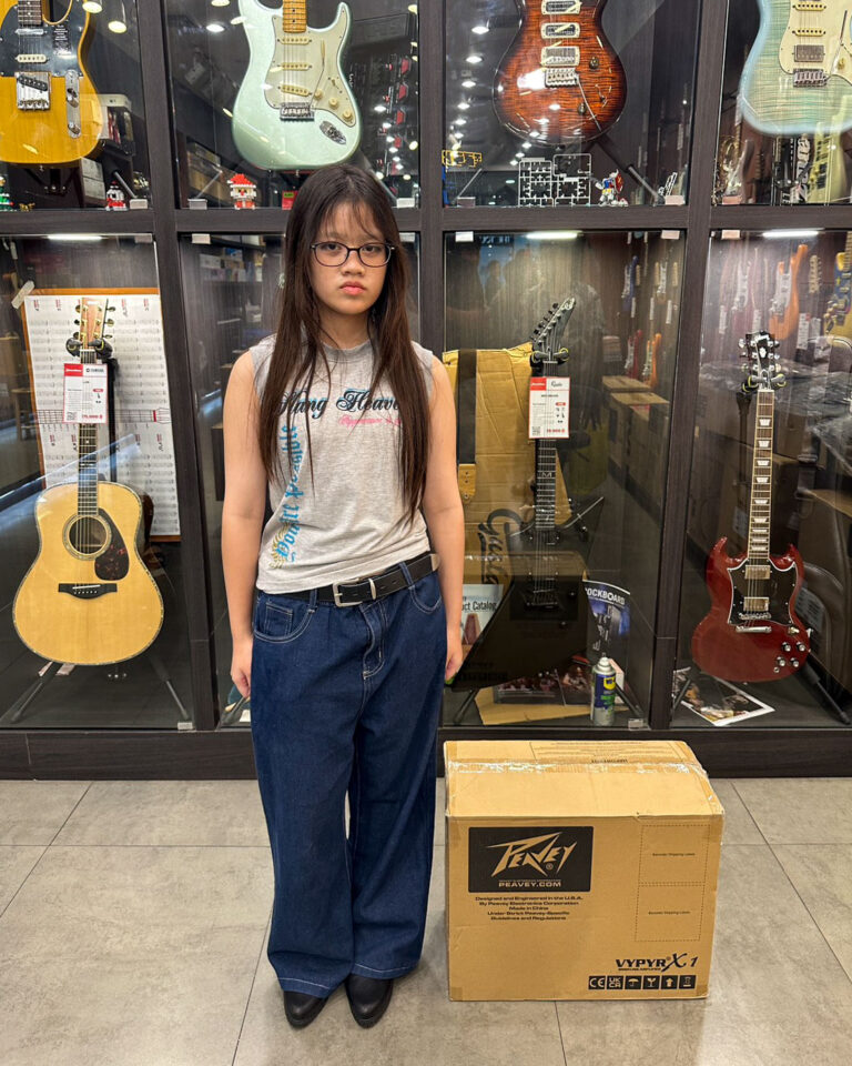 Girl wearing glasses standing in a guitar shop in front of glass display cases with guitars, a cardboard Peavey box at her side.