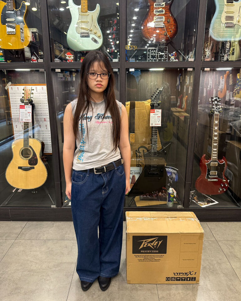 Girl standing in a guitar shop, with glass display cases of guitars behind her and a large Peavey box on the floor beside her.