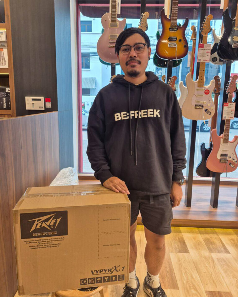 Man with glasses and goatee in a black hoodie stands beside a large Peavey box in a guitar shop with multiple electric guitars on display behind him. He rests one hand on the box.