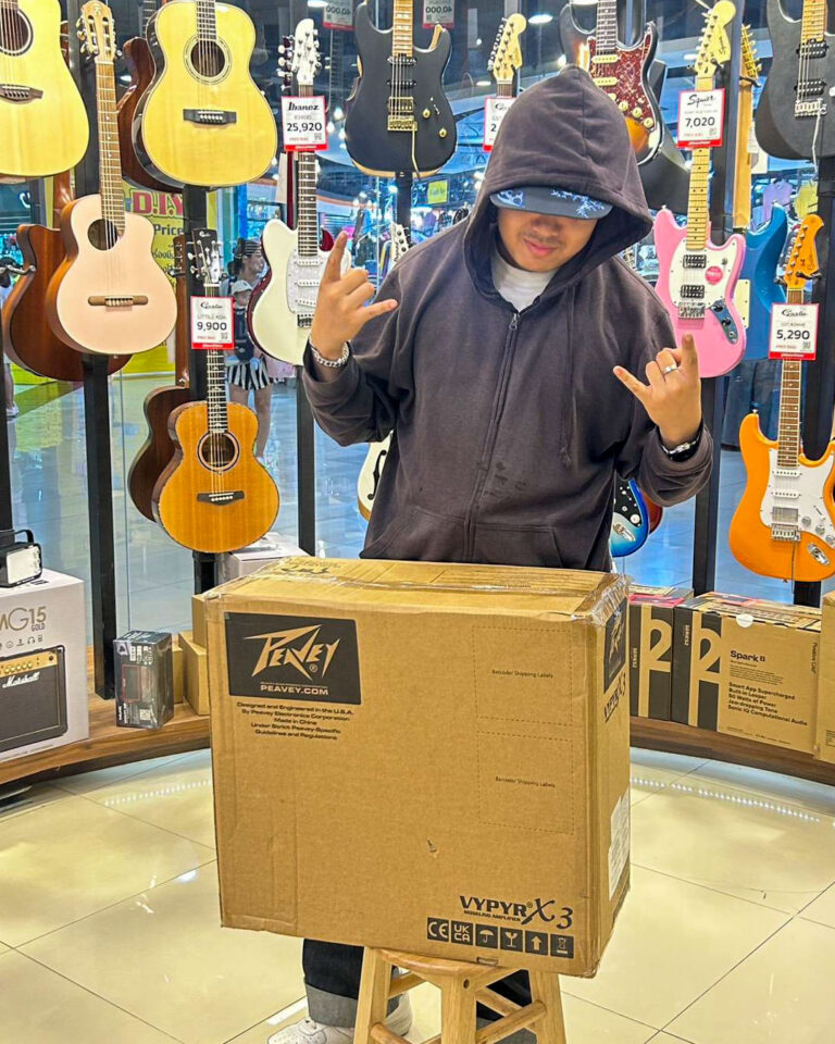 Person wearing a dark hoodie posing with hands raised in a rock gesture in a guitar shop, large cardboard box in front on a wooden stool.