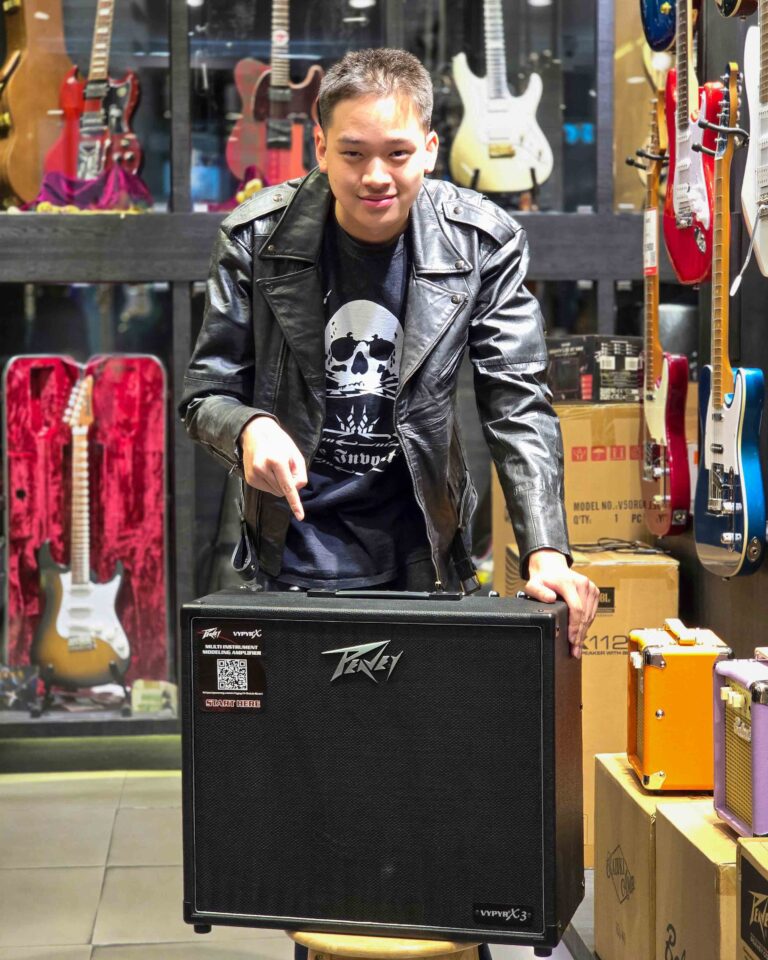 Man in a black leather jacket stands behind a large Peavey VypyrX3 guitar amplifier in a music store, with guitars on display in the background.