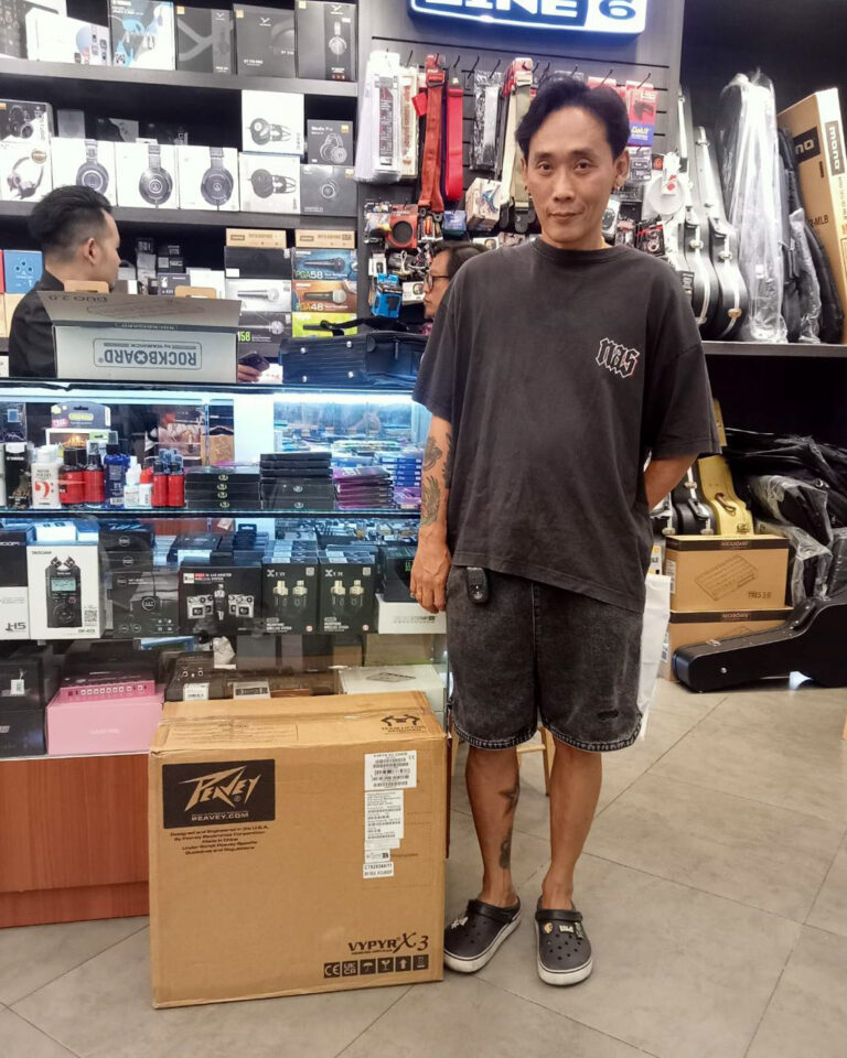 Man standing in a music store beside a large brown packing box labeled Peavey on the floor next to shelves of audio gear and guitars.