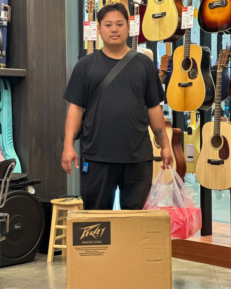 Man in a black t-shirt stands in a guitar shop, with several acoustic guitars hanging on the wall behind him and a plastic bag in his hand.