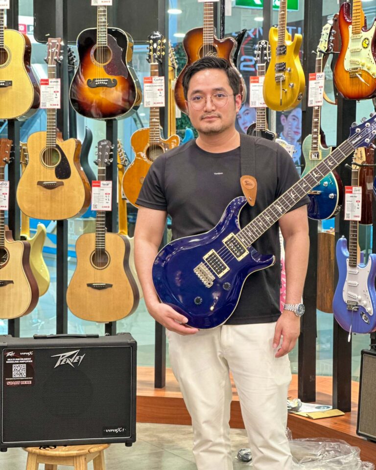 Man wearing a black t-shirt and glasses posing with a blue electric guitar in a guitar shop with guitars hanging on the wall behind him.
