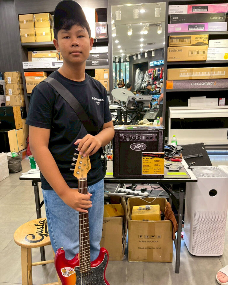 Young person in a black shirt and cap stands in a music shop holding a red electric guitar with a strap over the shoulder.