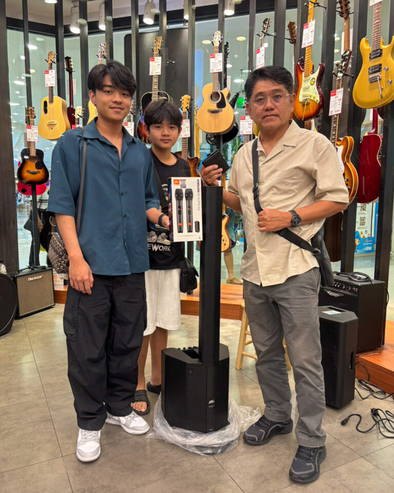 Three people in a guitar shop pose with a boxed wireless microphone kit on a speaker stand.