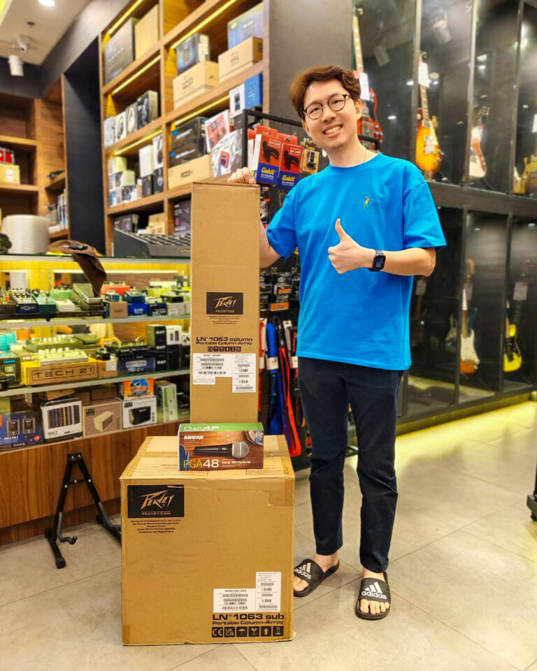 Smiling man in a blue shirt gives a thumbs-up beside stacked boxes in a music shop display area.
