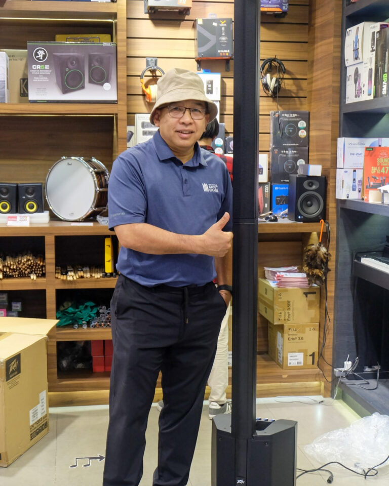 Man in a blue polo shirt and beige bucket hat giving a thumbs-up in an audio equipment shop.