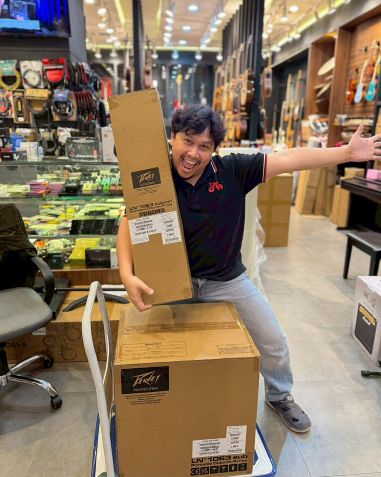 Happy man in a music store poses with a tall cardboard box on a hand truck, arms open wide.
