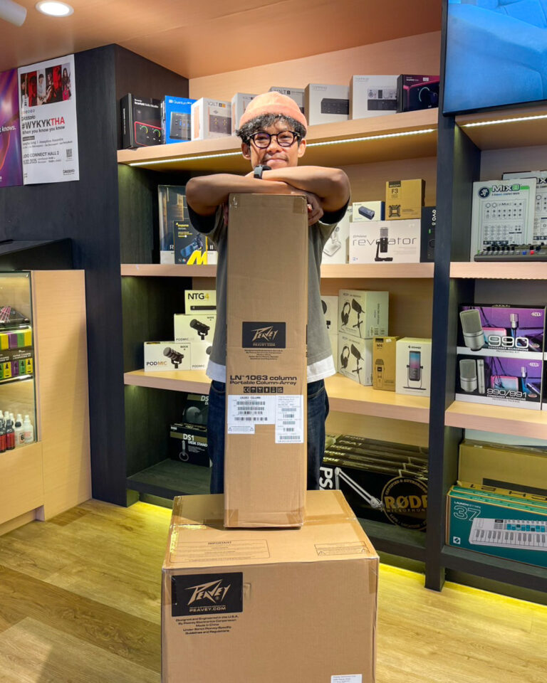 Young man wearing an orange beanie and glasses leans on a tall cardboard box in a tech shop, with shelves of electronics in the background.