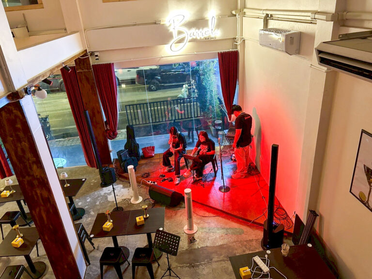 Intimate performance space with three musicians rehearsing on a red-lit stage beside a glass wall facing the street. Two players sit with instruments while a person adjusts equipment nearby.