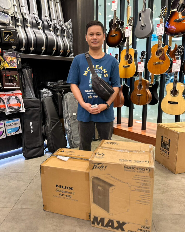 Man stands in a guitar shop beside boxed amplifiers, with acoustic guitars on display behind him.