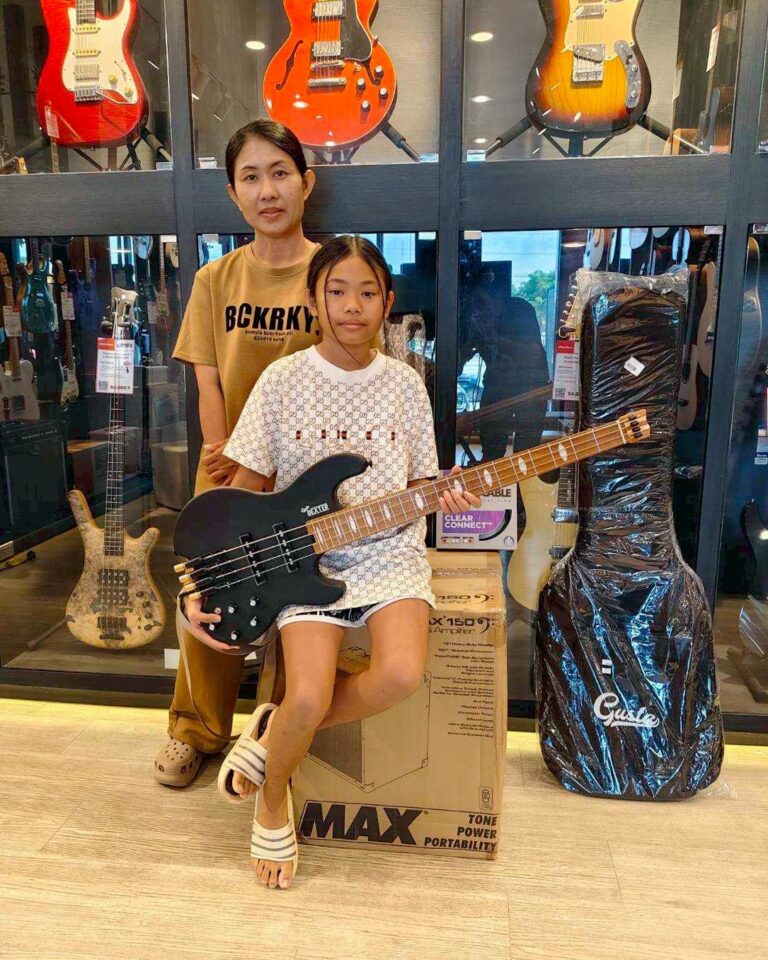 Girl sitting on a box holding a black bass guitar in a guitar shop, with an adult standing behind them among display guitars.