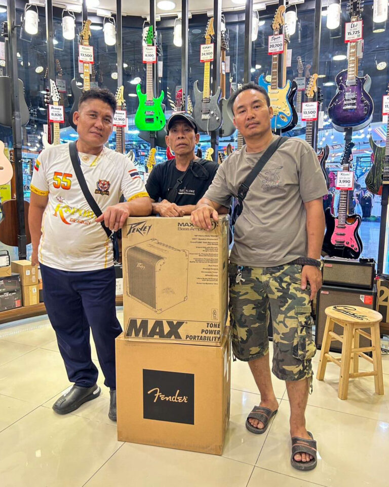 Three men stand in a guitar shop posing beside a large Fender amplifier box, with guitars hanging on the wall behind them.