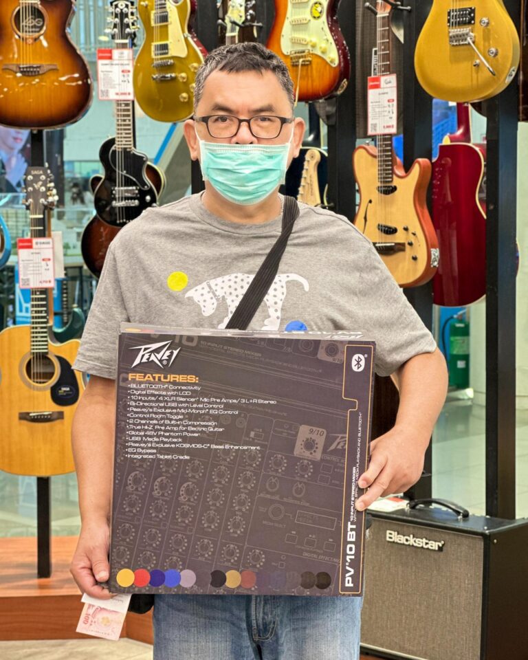Man wearing glasses and a face mask stands in a guitar shop, holding a large boxed audio product (Peavey) with guitars on display in the background.