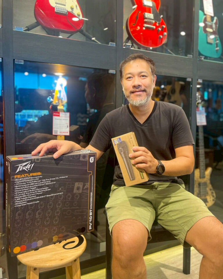 Smiling man in a black shirt and khaki shorts sits in a guitar shop, holding two boxed audio gear with guitar models visible behind him in glass displays.