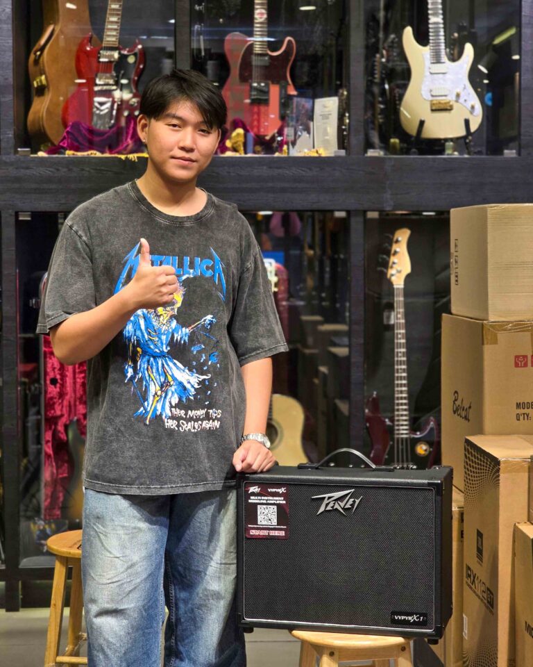 Young man in a dark graphic tee giving a thumbs up beside a guitar amplifier in a music store with guitars on display behind him.