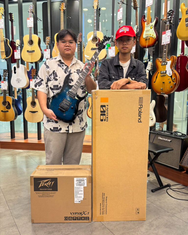 Two young men pose in a guitar shop, one holding a blue electric guitar while the other leans on a large Roland box, with many guitars on display behind them.
