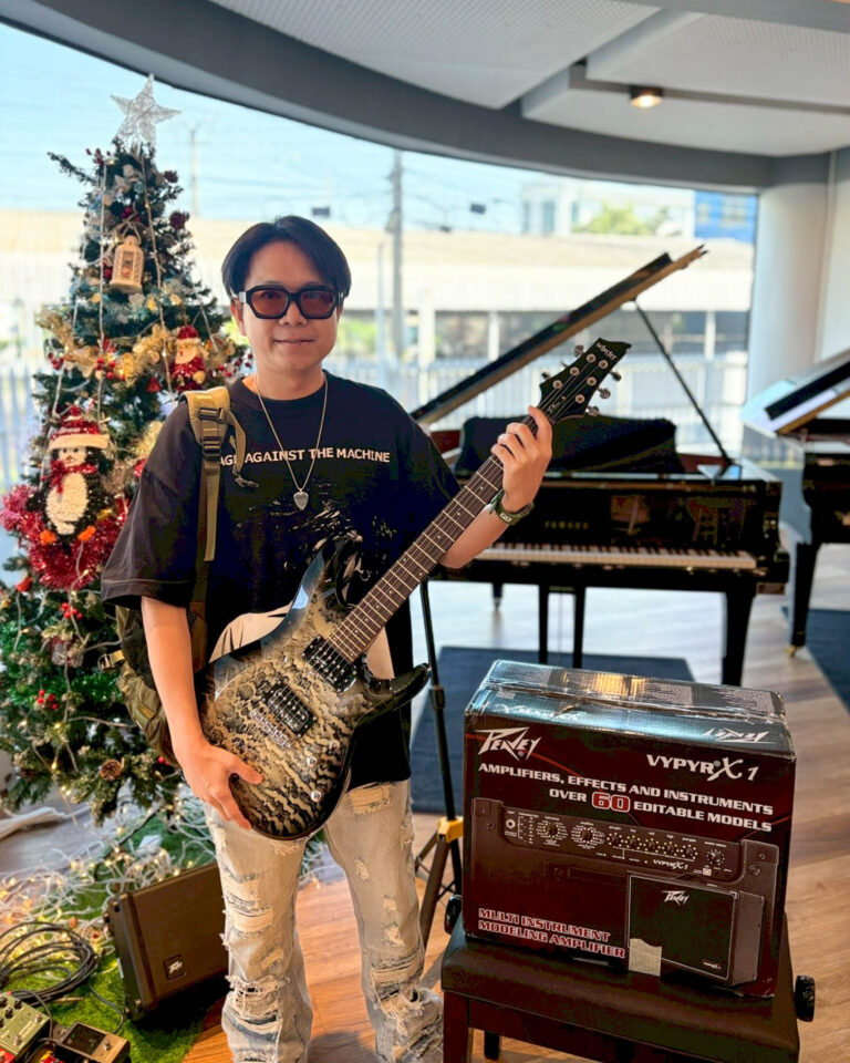 Young man with sunglasses holds a patterned electric guitar in a room with a Christmas tree and a Peavey amplifier box nearby; festive decor visible in background.