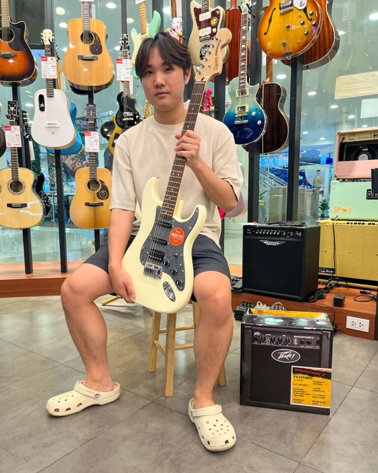 Young man sits on a stool in a guitar shop, holding a white electric guitar among wall-lined guitars.EDt