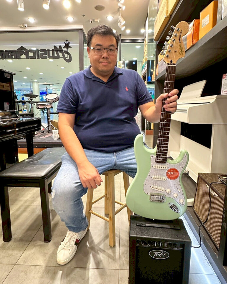 Man in a navy polo sits on a wooden stool, holding a mint-green Fender Stratocaster in a music store with a piano and drums in the background.