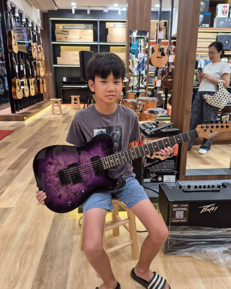 Young boy sitting on a stool in a guitar shop, holding a purple electric guitar with a brick wall of instruments in the background.