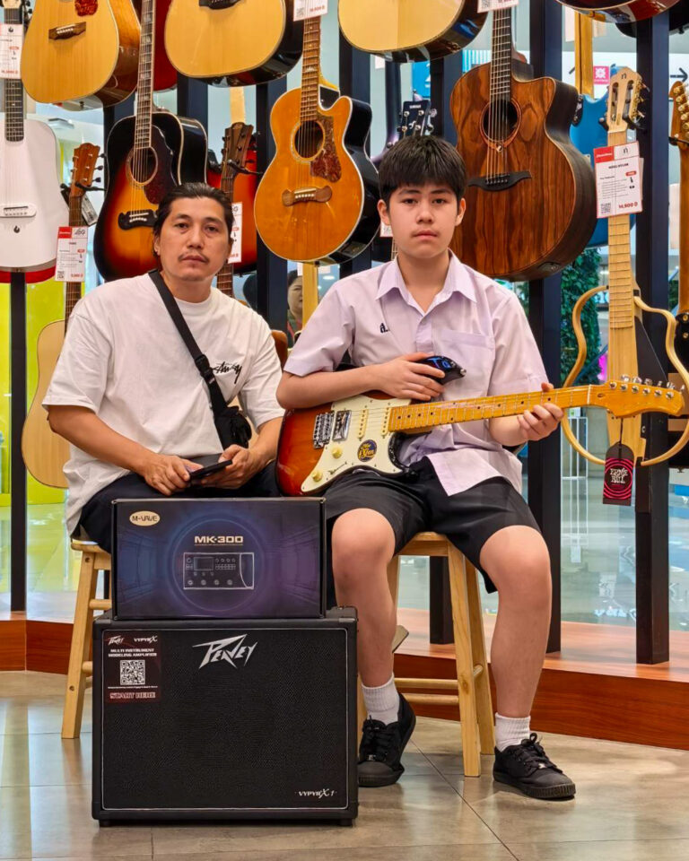 Two people sit in a guitar shop surrounded by hanging guitars; the boy holds an electric guitar while the man beside him holds a phone.