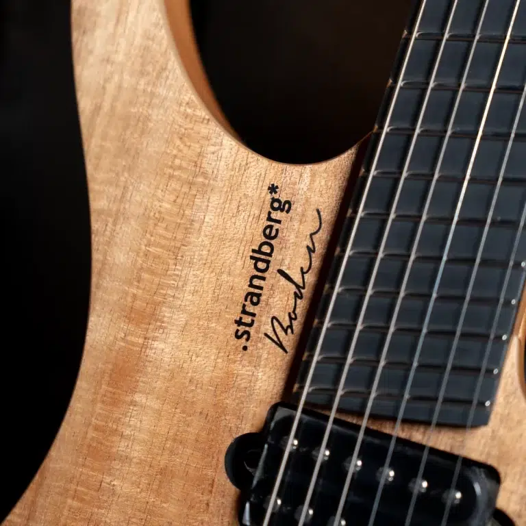 Close-up of a natural wood Strandberg electric guitar neck, showing the logo and a handwritten signature along the fretboard with strings nearby. ขายราคาพิเศษ