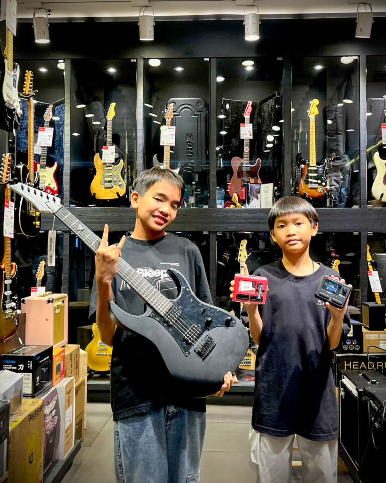 Two boys stand in a guitar shop; one holds a black electric guitar and points upward with a hand gesture, the other holds guitar pedals.