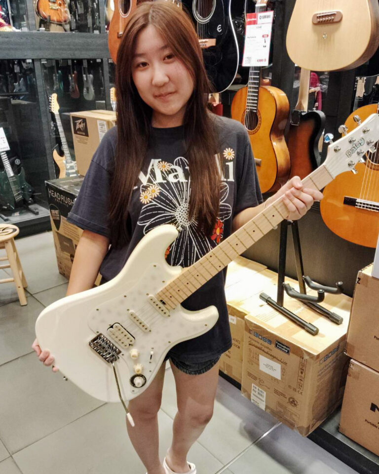 Young girl in a dark graphic shirt holds a white electric guitar in a guitar shop, surrounded by instruments on display.