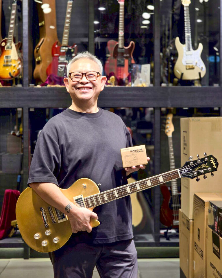Smiling man in a guitar shop holds a gold electric guitar and a small cardboard box in front of a wall of guitars behind him.