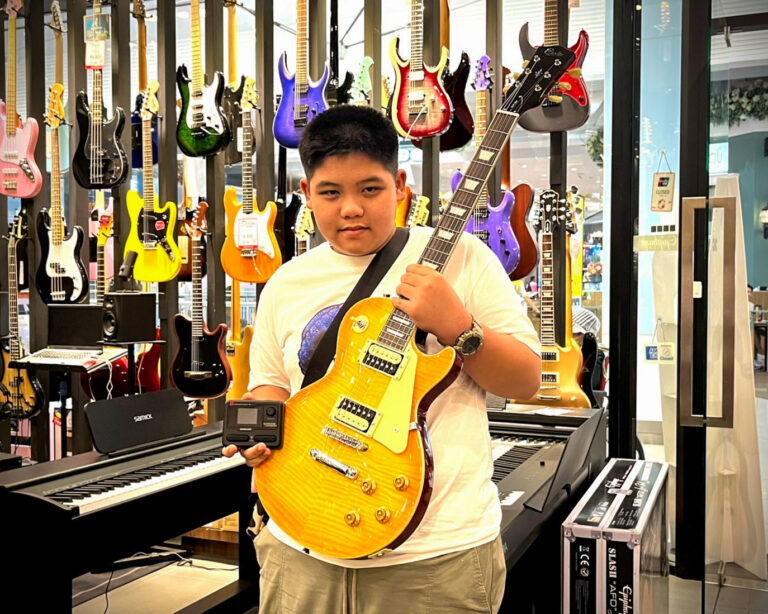 Young person in a guitar shop posing with a sunburst yellow electric guitar amid rows of colorful guitars on display behind him.