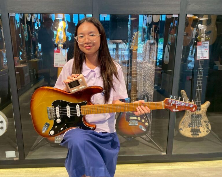 Young girl with clear glasses poses with a sunburst electric guitar in a glass-display guitar shop.