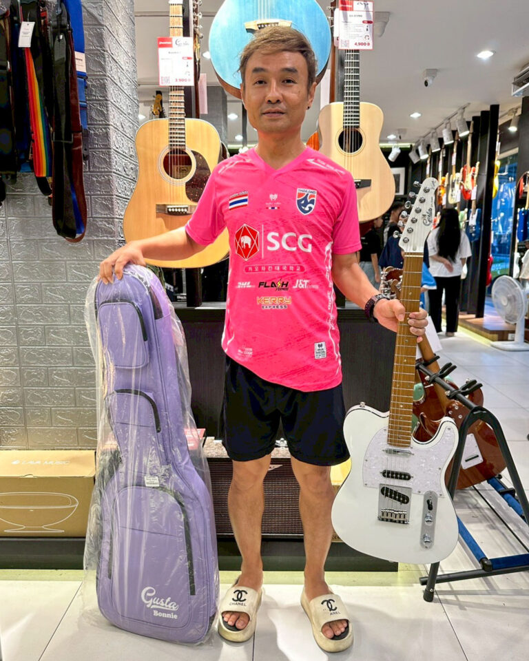Man in a pink sports jersey holds a white electric guitar in a guitar shop, with a purple guitar case beside him.