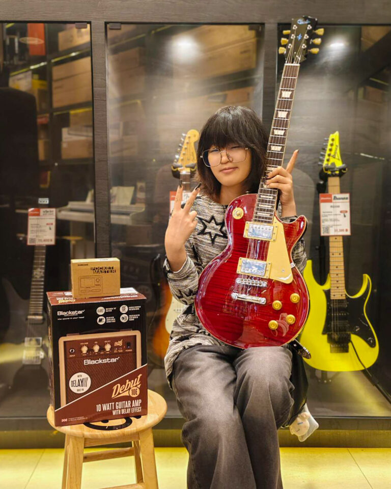 Person sitting in a guitar shop, holding a red electric guitar and flashing a peace sign to the camera.